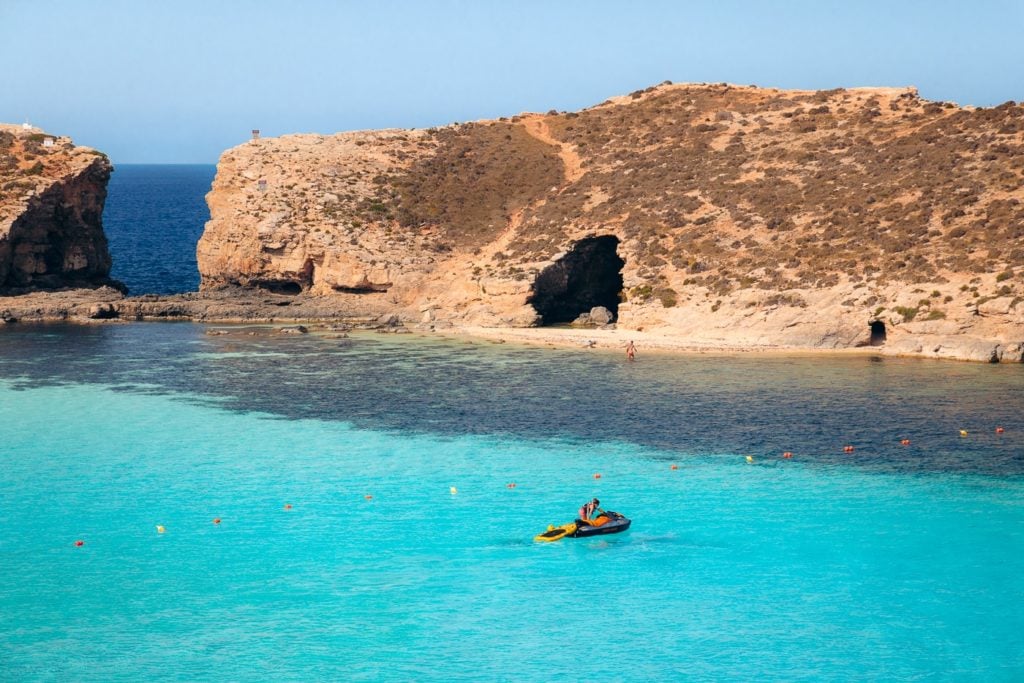Cominotto Island, Malta, featuring crystal-clear turquoise waters, rugged cliffs, and a serene beach, ideal for private boat tours.