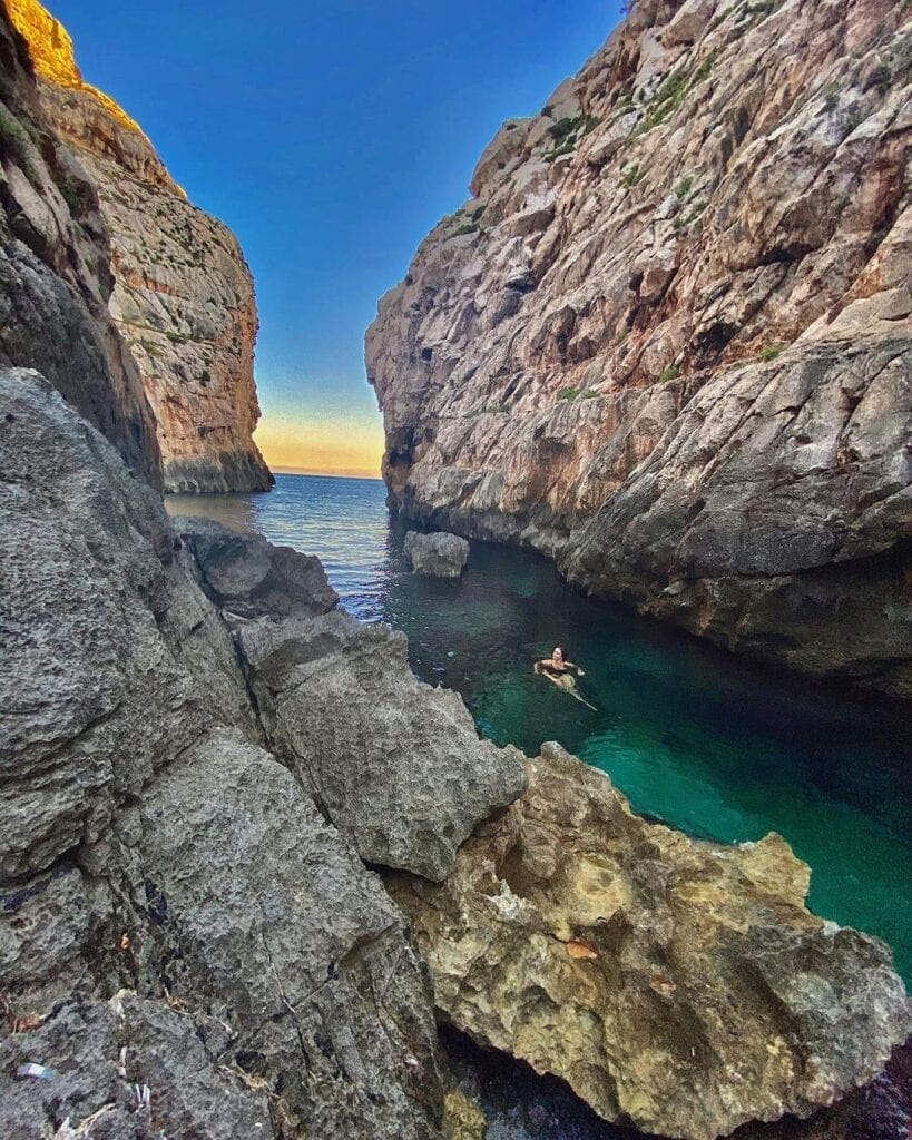 Crystal-clear blue waters at Wied iz-Zurrieq, Malta, with swimmers enjoying a refreshing dip near the rugged limestone cliffs.