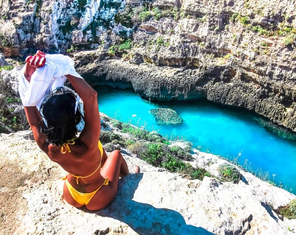 Woman in a bikini sitting by the turquoise waters of Wied il-Għasri, Gozo, surrounded by dramatic limestone cliffs and a secluded rocky beach.