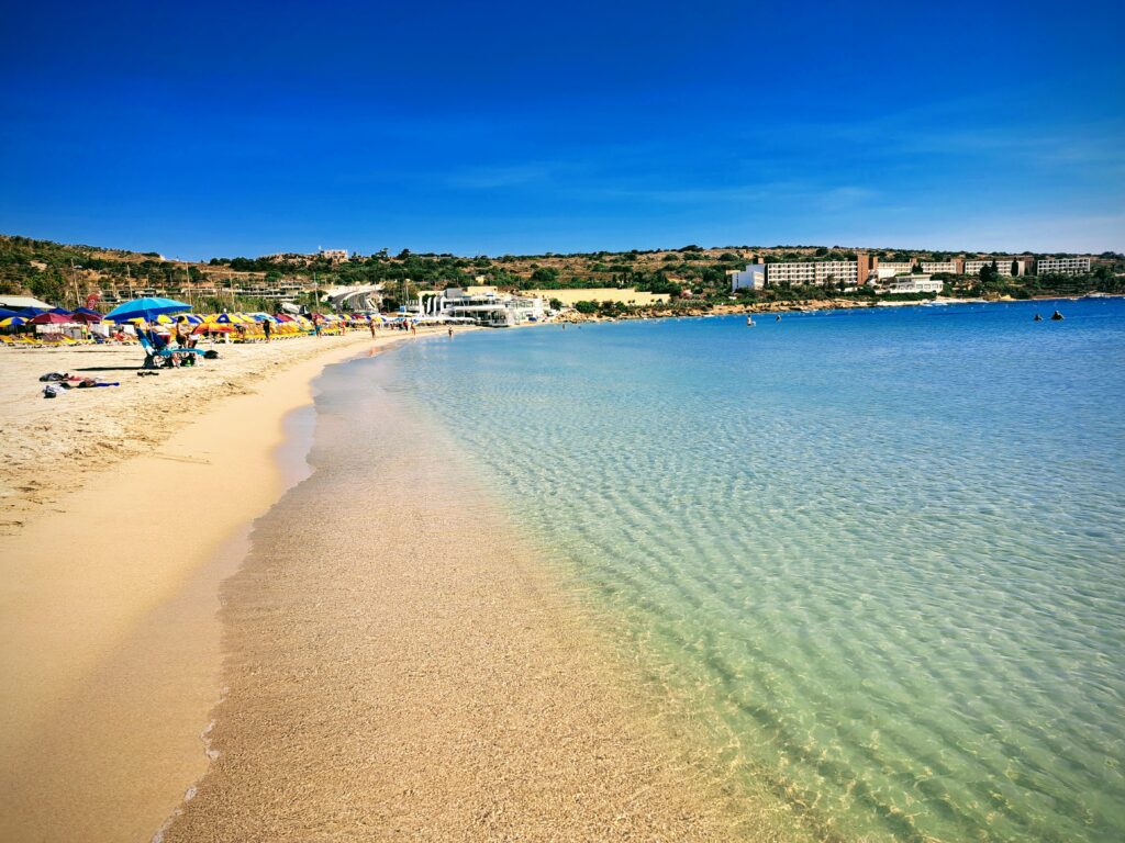 Panoramic view of Mellieħa Bay, Malta, featuring a wide sandy beach, calm turquoise waters, and a backdrop of rolling hills and charming seaside buildings