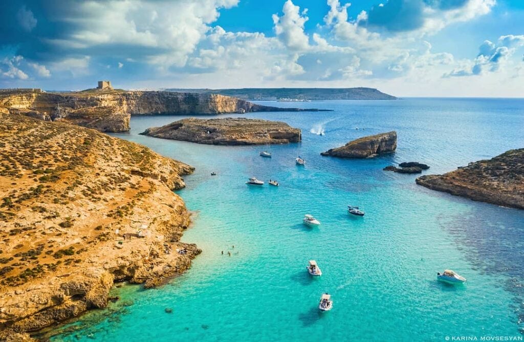 Crystal-clear turquoise waters of the Blue Lagoon, Malta, surrounded by rugged limestone cliffs and bathed in golden sunlight, with boats anchored nearby and swimmers enjoying the serene paradise.