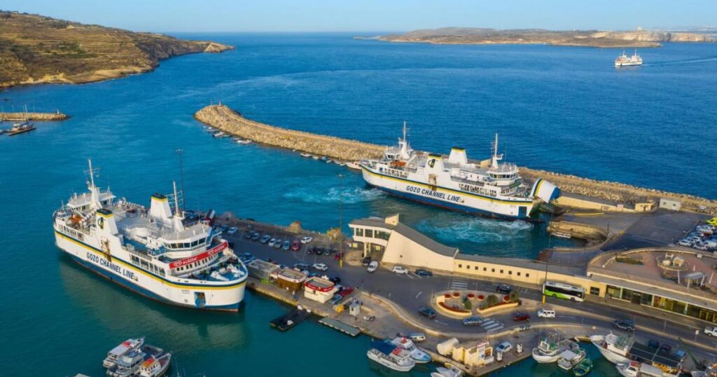 Gozo Channel ferry docked at the terminal, with vehicles boarding and the coastline visible in the background.