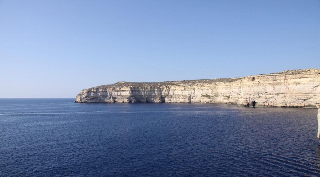 Scenic view of Ghar Qawqla Bay in Gozo, Malta, featuring crystal-clear turquoise waters, rugged limestone formations, and a serene coastal landscape under a bright blue sky