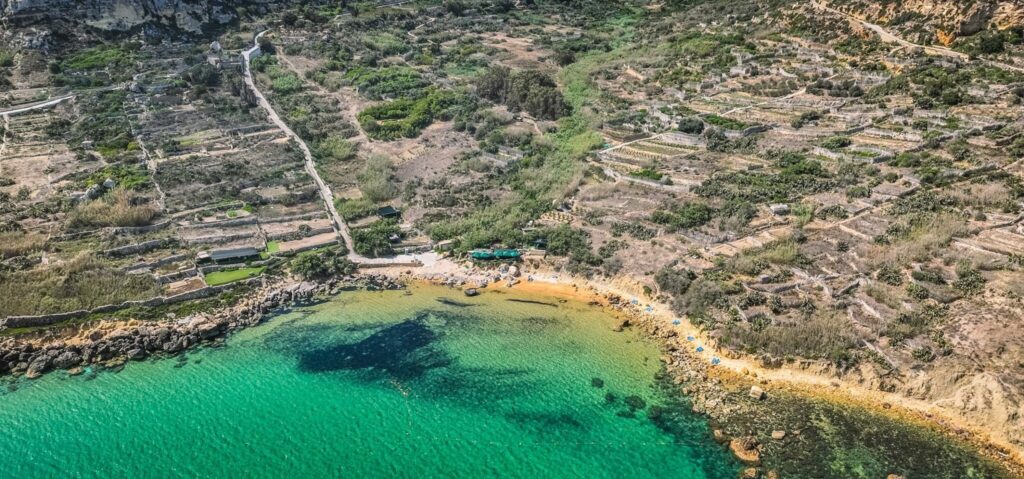 Scenic view of San Blas Beach in Malta, featuring golden-red sand, turquoise waters, and lush green hills in the background. The secluded cove is surrounded by rugged cliffs, with a few beachgoers enjoying the tranquil atmosphere.