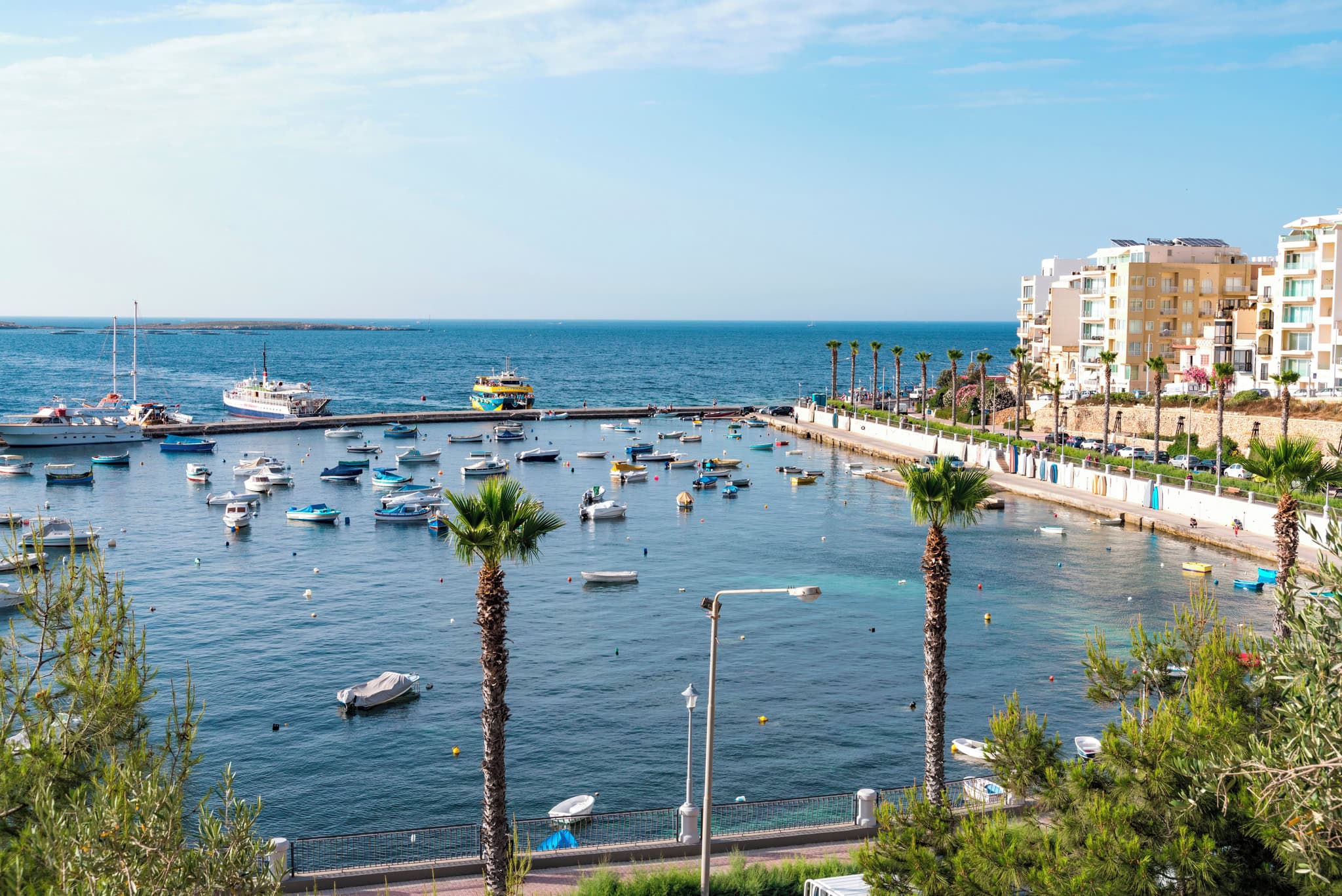 Picturesque view of St Paul’s Bay Malta with calm blue waters, fishing boats, waterfront promenade, and coastal buildings under a clear sky.