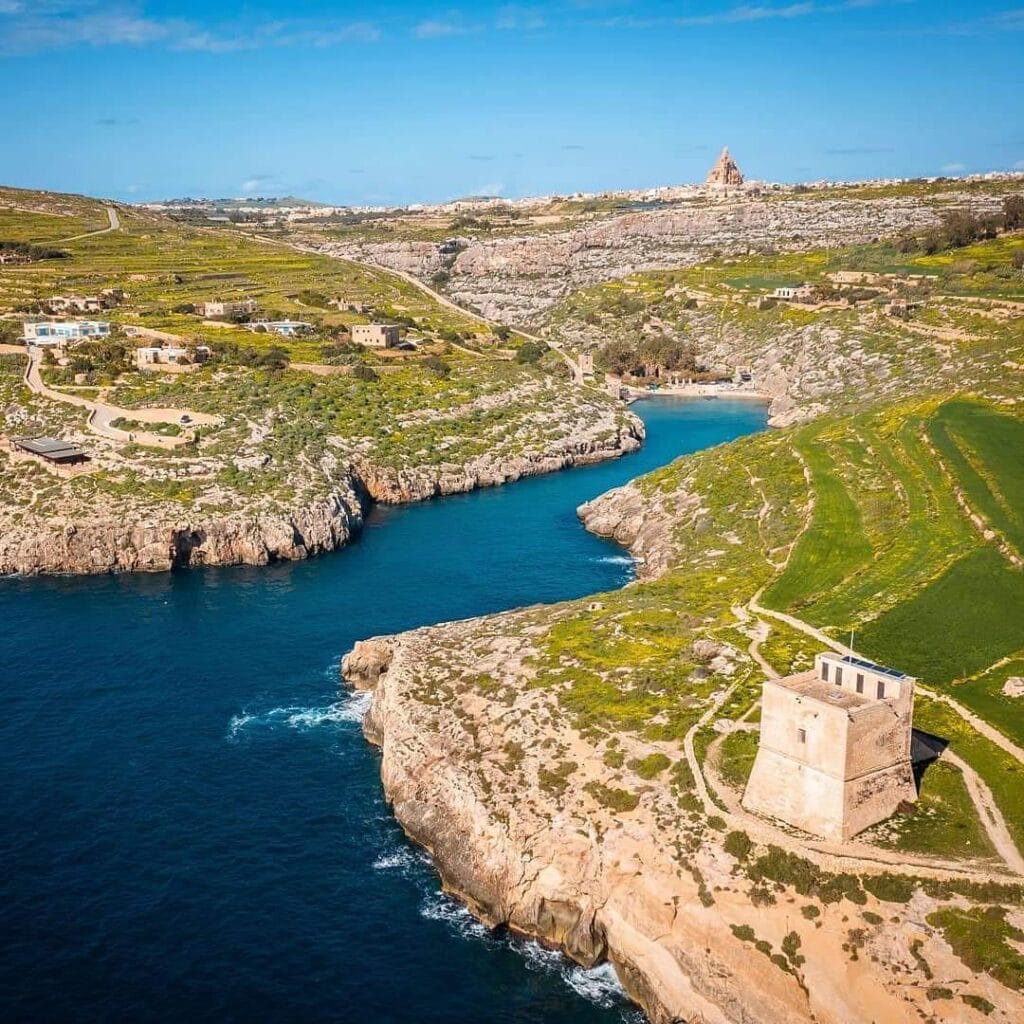 A wide shot of the Mġarr ix-Xini bay from the water, showing the cliffs on both sides and the tower in the distance.