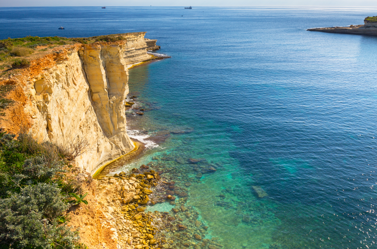 View of the rocky cliffs near Marsaxlokk Fishing Village, Malta, with the sea stretching into the horizon and traditional Maltese fishing boats in the distance.