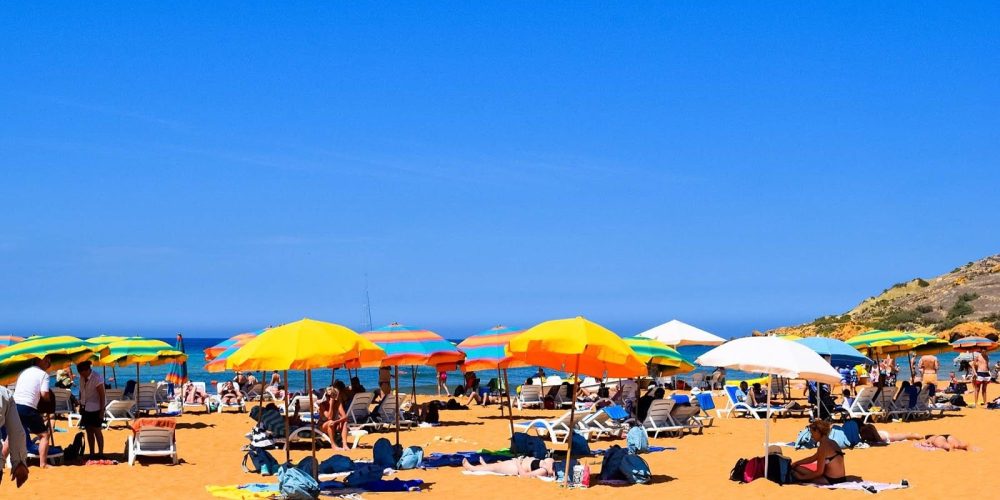 Ramla Bay Scenic view of Ramla Bay with golden sand, clear blue waters, and tourists relaxing under umbrellas on a sunny day.