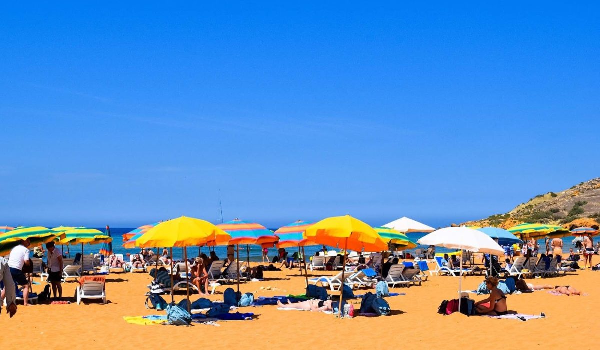 Scenic view of Ramla Bay with golden sand, clear blue waters, and tourists relaxing under umbrellas on a sunny day.