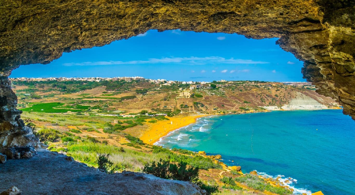 San Blas Beach in Malta Scenic view of San Blas Beach in Malta, featuring golden-red sand, turquoise waters, and lush green cliffs
