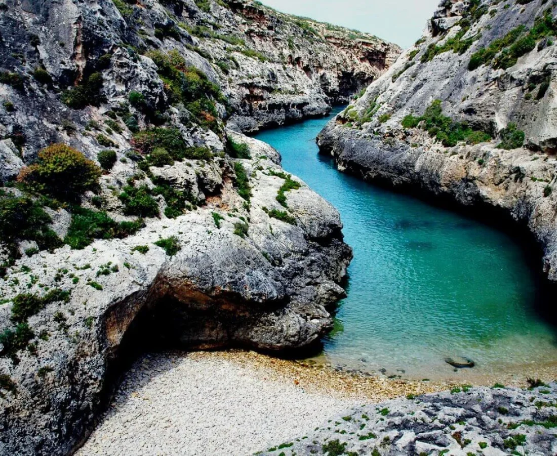 Scenic view of Wied il-Għasri, a narrow gorge in Gozo, Malta, featuring turquoise waters flowing between towering limestone cliffs, with a small pebble beach at the inlet.