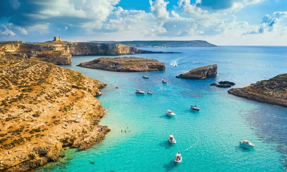 Crystal-clear turquoise waters of the Blue Lagoon, Malta, surrounded by rugged limestone cliffs and bathed in golden sunlight, with boats anchored nearby and swimmers enjoying the serene paradise.