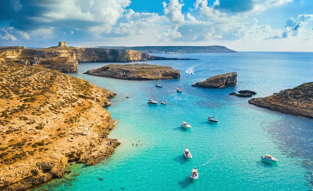 blue lagoon malta Crystal-clear turquoise waters of the Blue Lagoon, Malta, surrounded by rugged limestone cliffs and bathed in golden sunlight, with boats anchored nearby and swimmers enjoying the serene paradise.