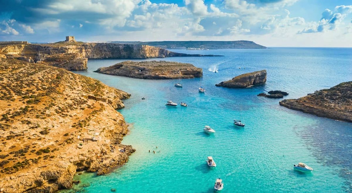 Crystal-clear turquoise waters of the Blue Lagoon, Malta, surrounded by rugged limestone cliffs and bathed in golden sunlight, with boats anchored nearby and swimmers enjoying the serene paradise.